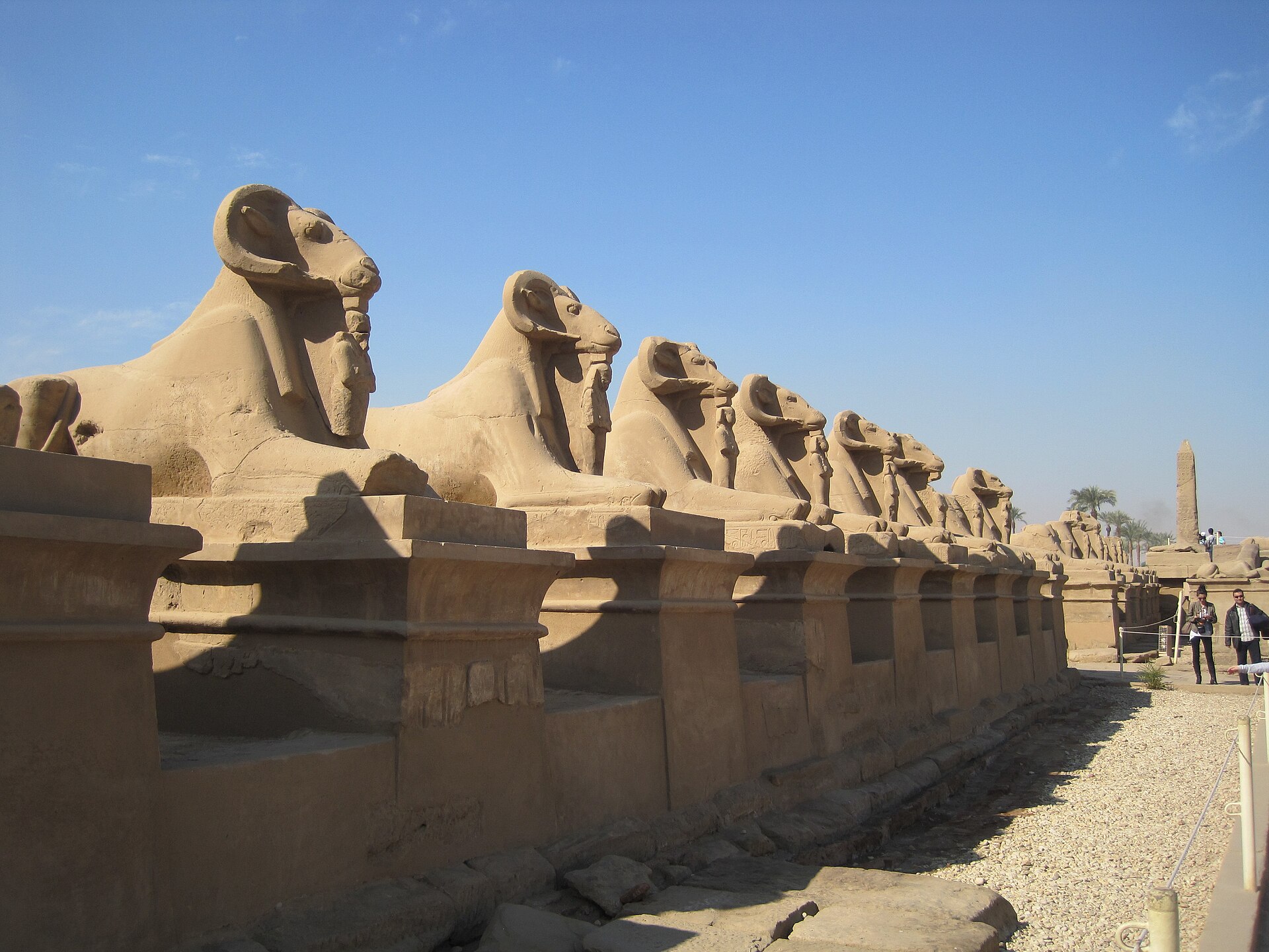 Close perspective down a line of ram-headed Karnak sphinxes on modern bases with an obelisk and visitors beyond.