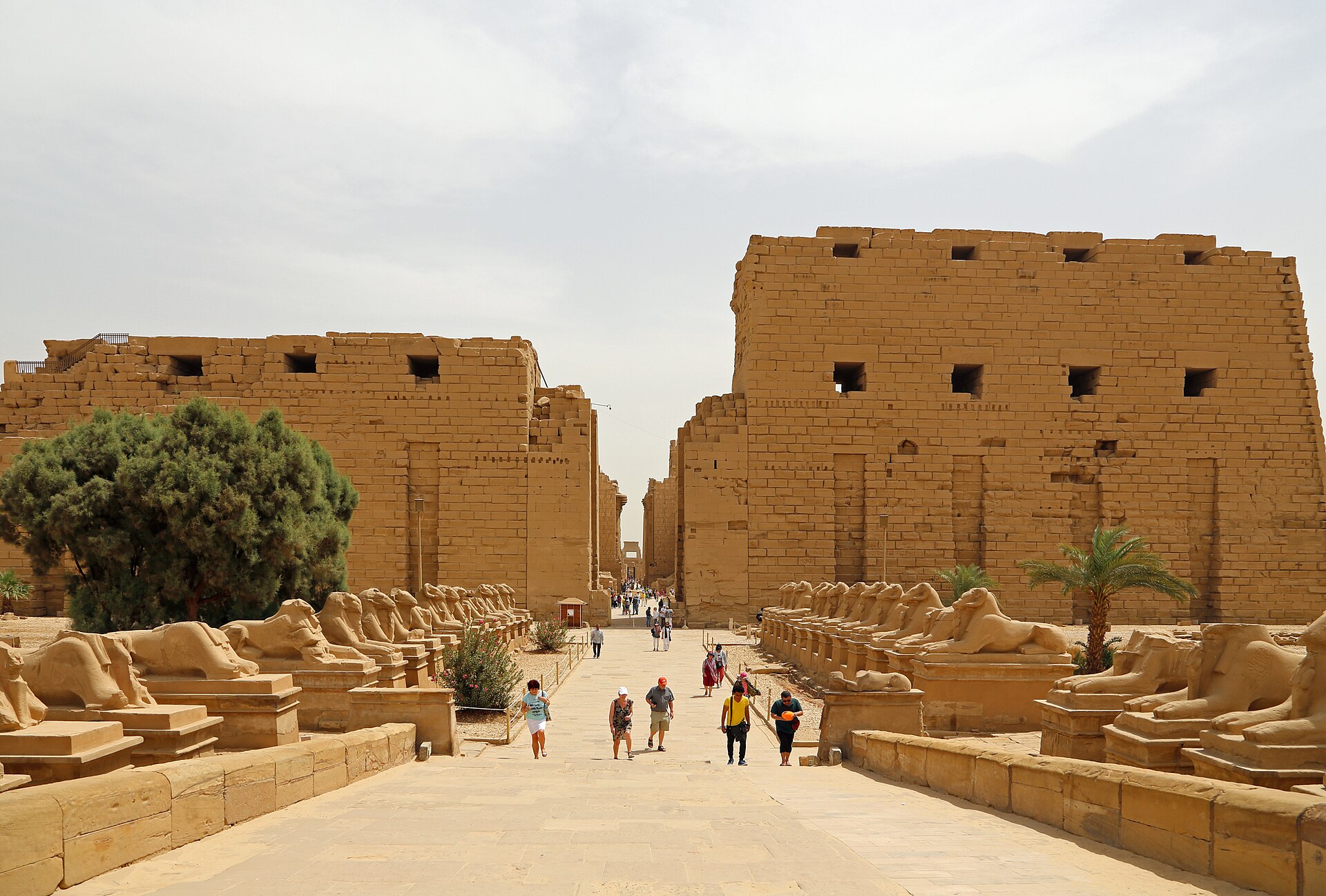 Unequal unfinished first-pylon towers behind the present ram-sphinx approach and visitors.