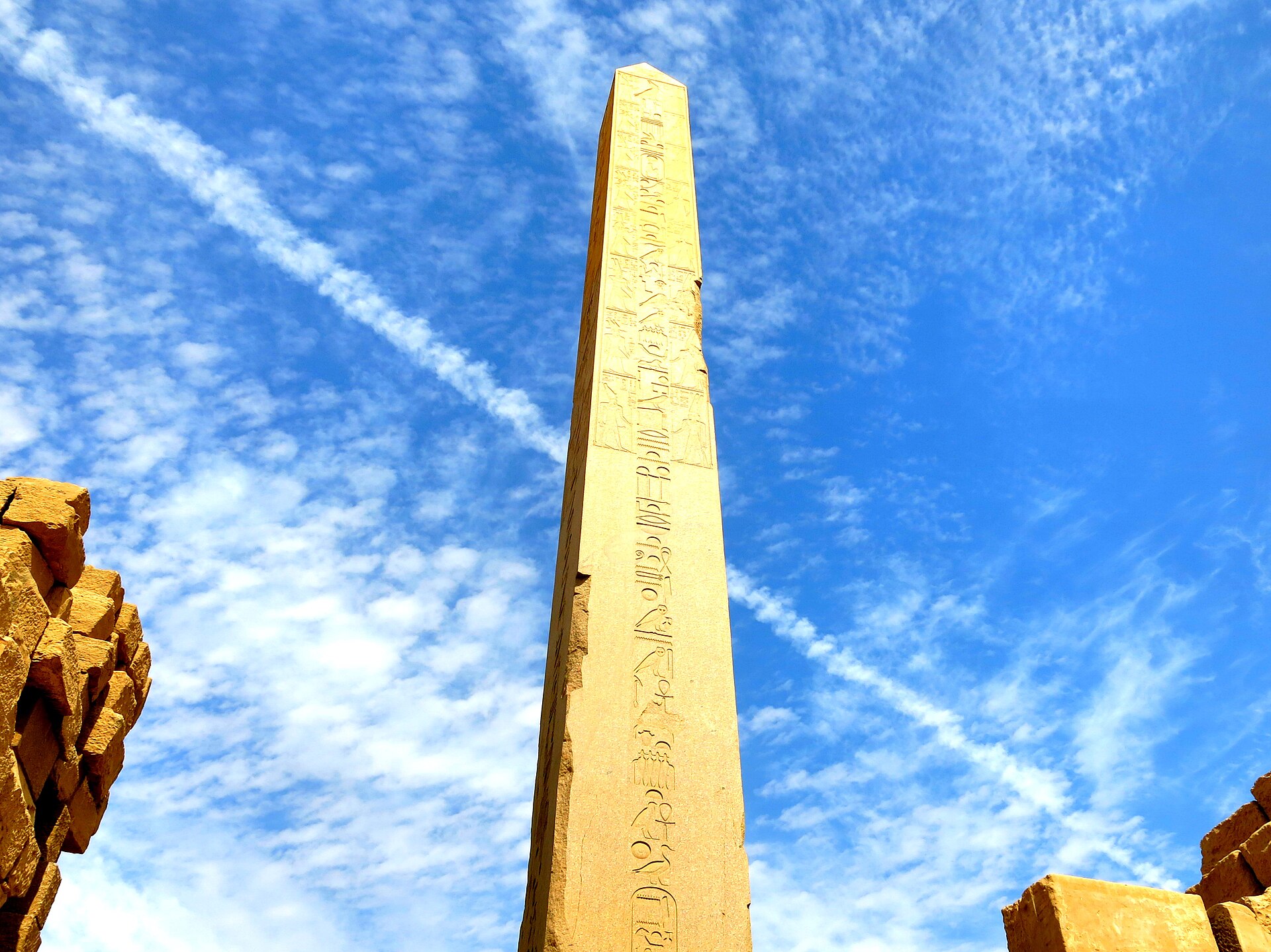 Tall granite obelisk of Hatshepsut rises from the Karnak ruins against a blue sky, with hieroglyphic inscriptions on its face.