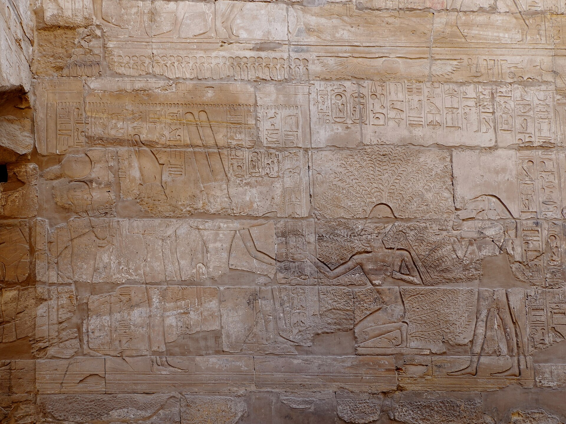 Worn royal and divine relief figures with hieroglyphic bands carved on a sandstone hall wall.