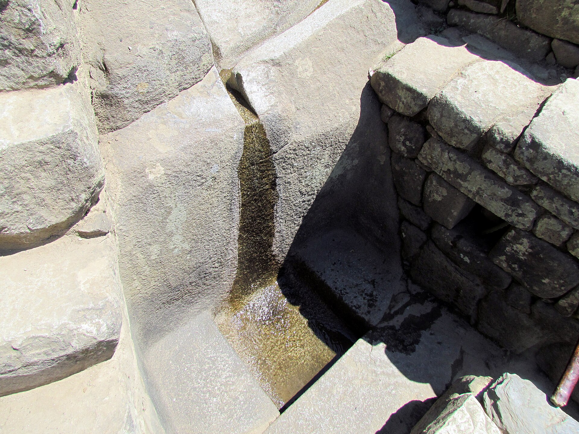Close overhead view of a narrow wet carved-stone channel and receiving basin beside fitted fountain-area masonry.