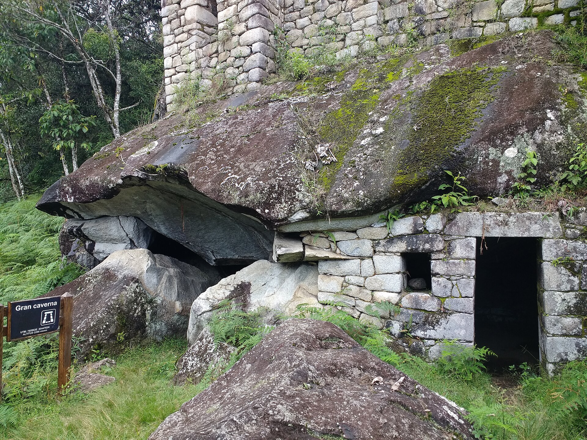Waynapicchu Gran Caverna rock cavity under a stone-built wall and opening, amid ferns and a modern visitor sign.