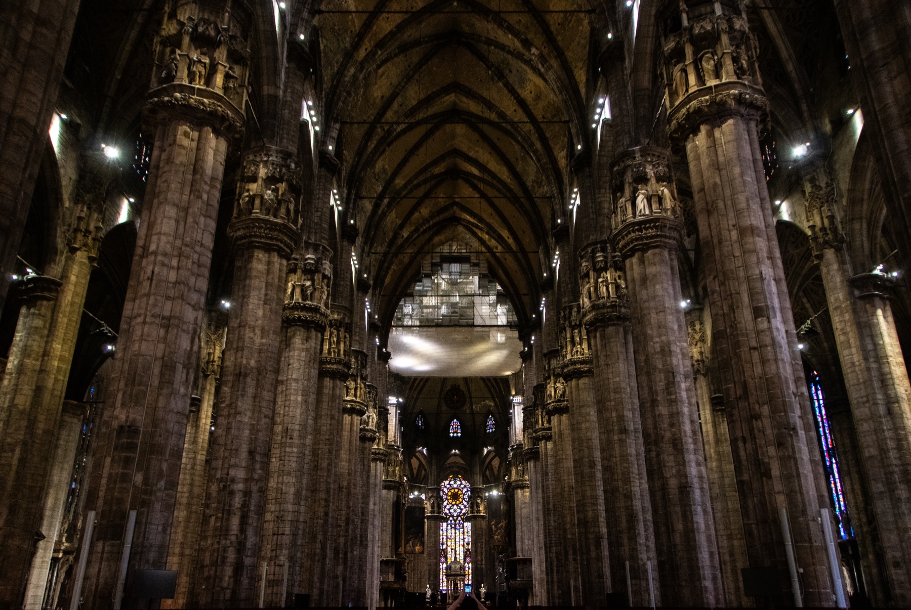 Milan Cathedral’s dark central nave with immense clustered piers, carved capitals, pointed rib vaults, iron ties and the glowing east end.