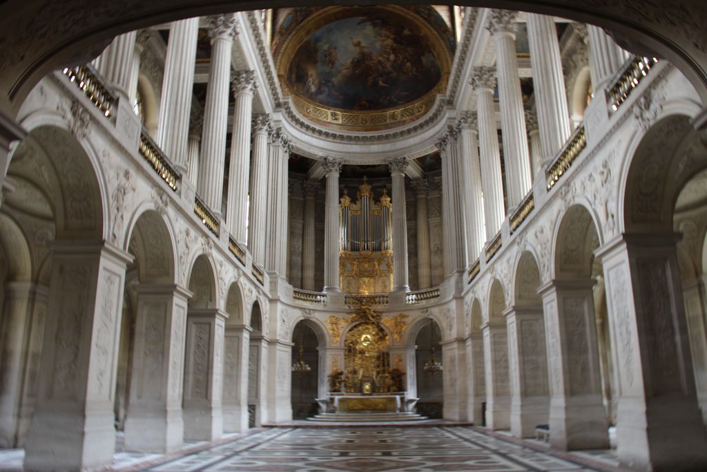A tall white-and-gold chapel has a lower arcade, upper colonnade, royal galleries, painted vault, organ and patterned marble floor.
