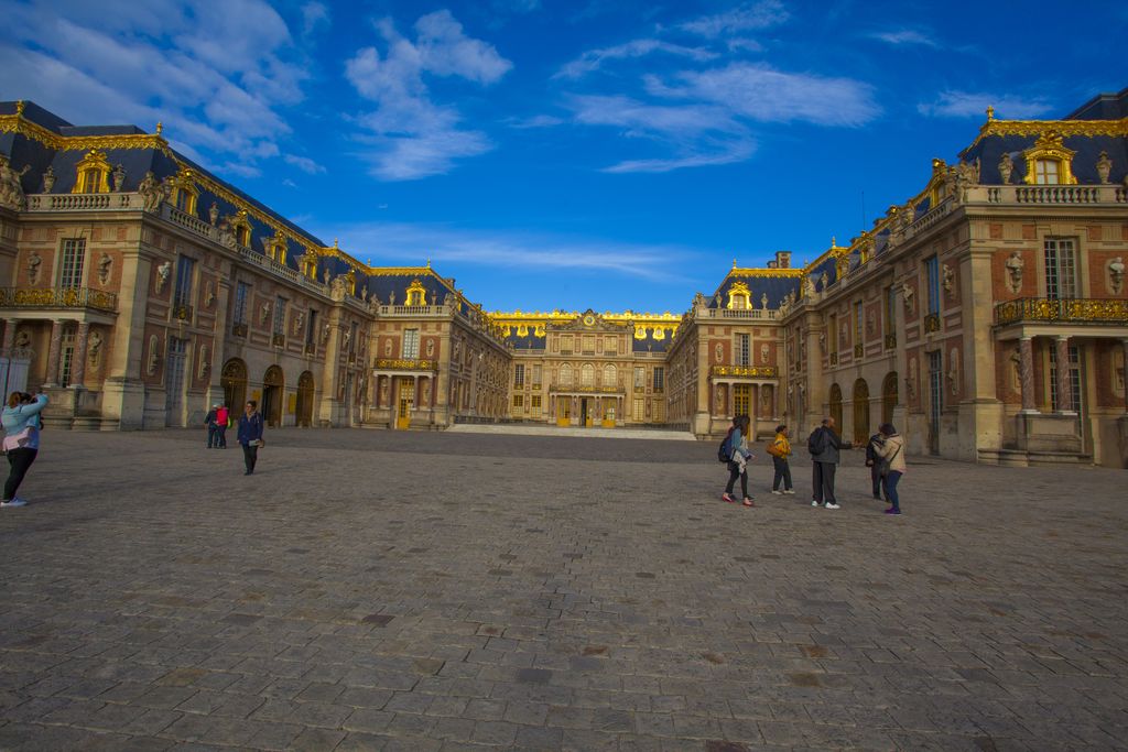 A broad paved court is enclosed by red-brick and pale-stone palace fronts with steep slate roofs, gilded balconies and scattered visitors.