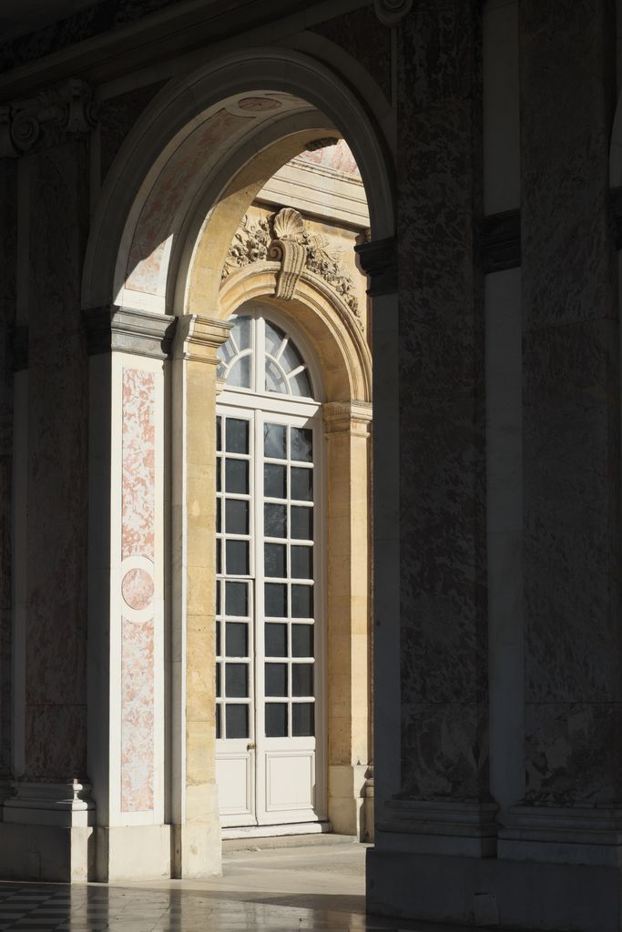 A tall arch lined with pink-veined marble opens from a shadowed peristyle toward a sunlit glazed door and courtyard wall.