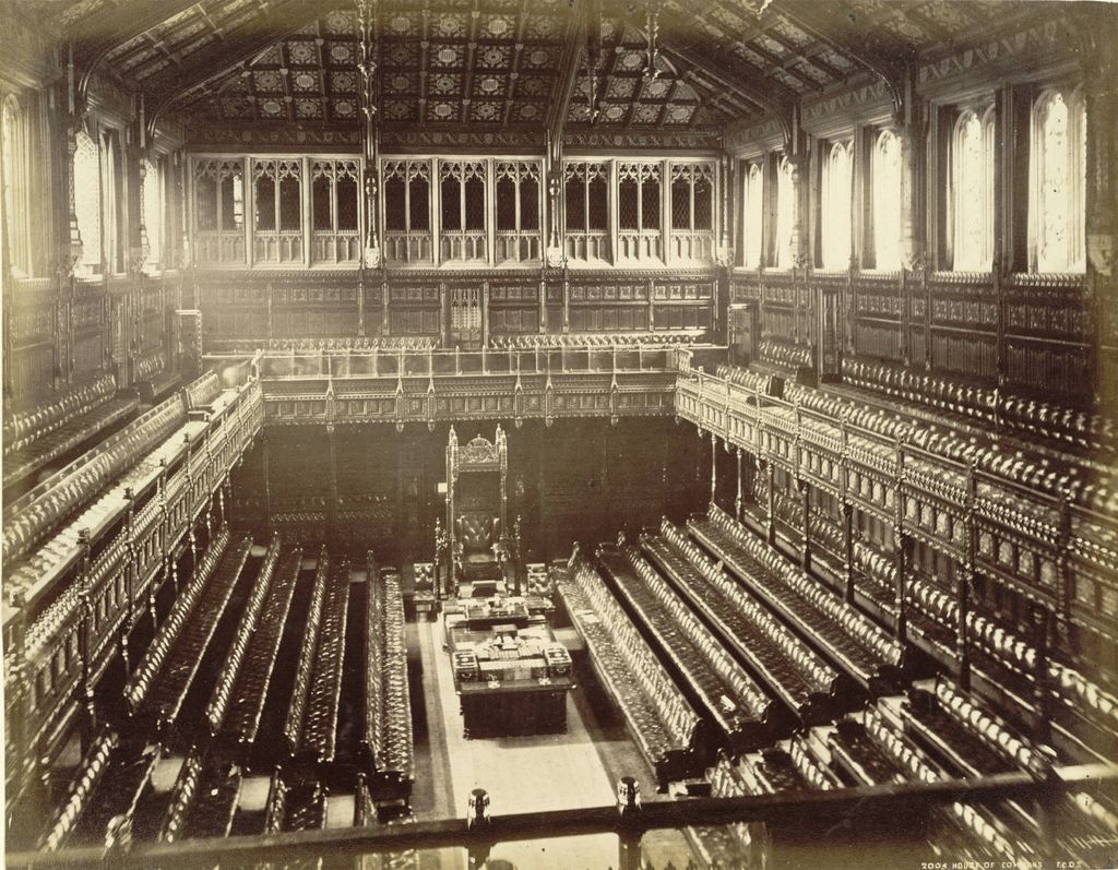 A sepia historical photograph looks down into a narrow parliamentary chamber lined by long facing benches beneath galleries and a patterned ceiling.