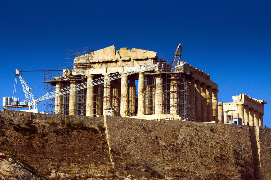 The Parthenon rises above a massive Acropolis wall under a deep blue sky, almost encased in scaffolding and crossed by two large conservation cranes.