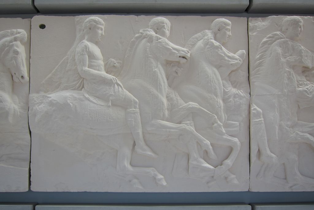 Pale rectangular relief blocks in a museum show overlapping horsemen, horses and standing figures, with vertical joints visible between the panels.