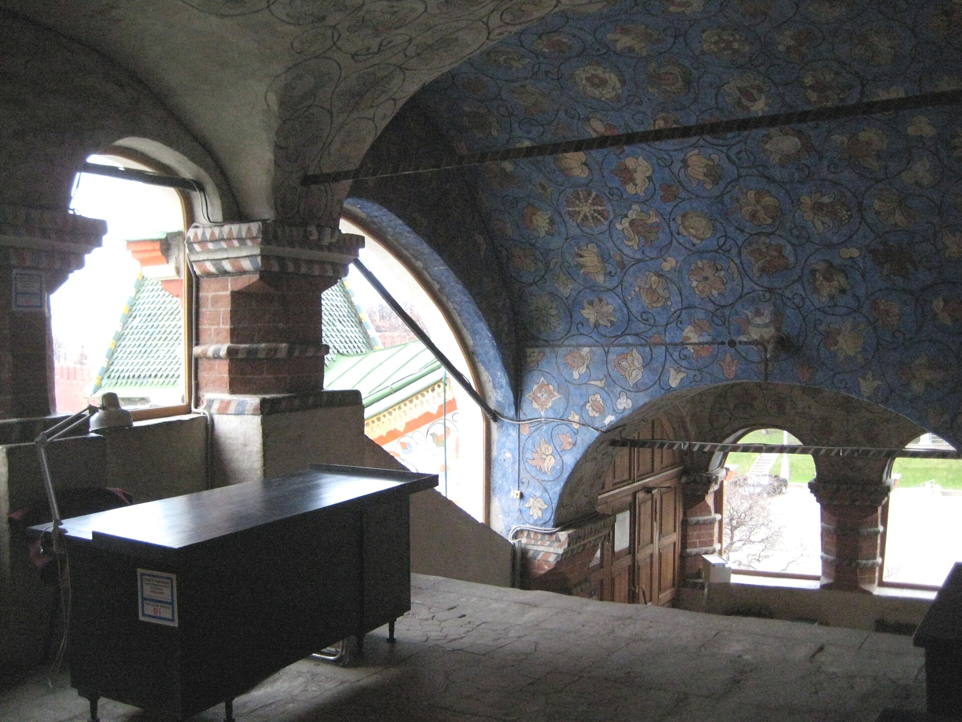 Enclosed gallery passage with curved openings, floral wall painting and views toward roofs outside.