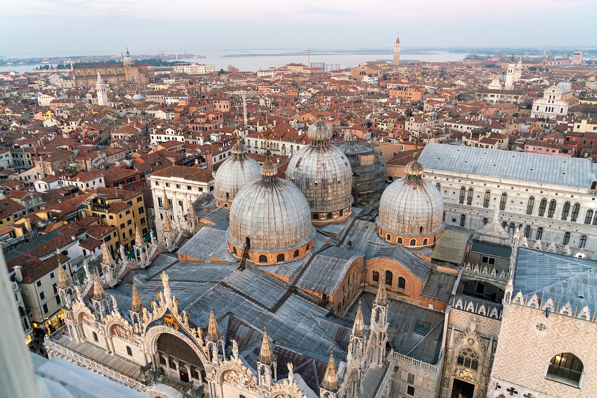 Elevated view over St Mark’s Basilica and the Doge’s Palace, showing lead-covered domes, brick roof structures and the surrounding lagoon city.