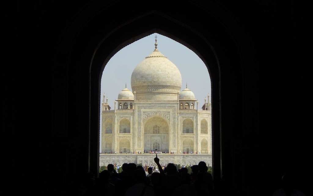 The pale dome and central façade of the Taj Mahal are framed by a deep black pointed gateway above a band of silhouetted visitors.