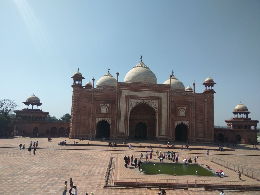 A broad red sandstone guest house has a large central pointed arch, three pale domes, white inlay and small domed pavilions around a paved court.