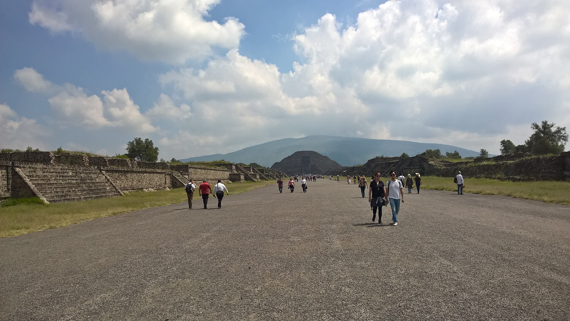Long straight visitor route with stepped ancient side structures, the Moon Pyramid and Cerro Gordo beyond.