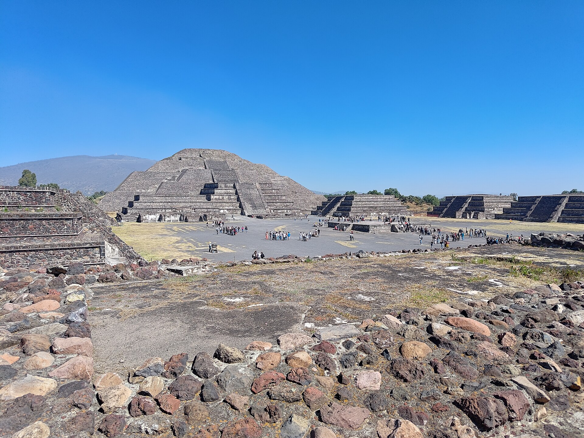 Moon Pyramid against Cerro Gordo above an open plaza and smaller stepped platforms, with modern visitors.