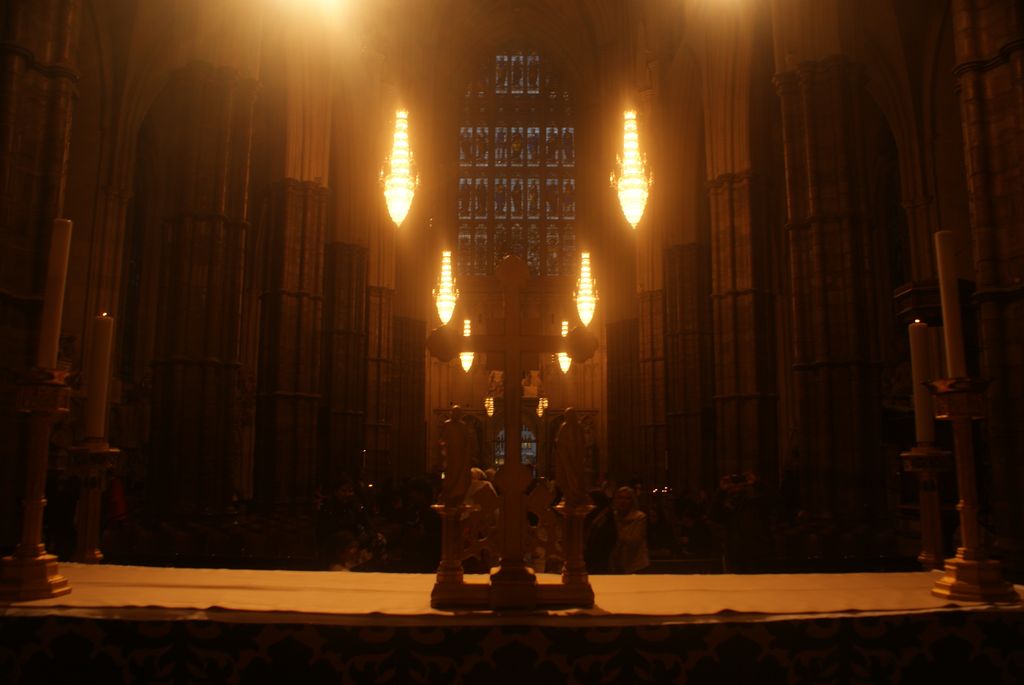 A dark symmetrical church interior has tall stone piers, glowing hanging lamps and a distant stained-glass window along its central axis.