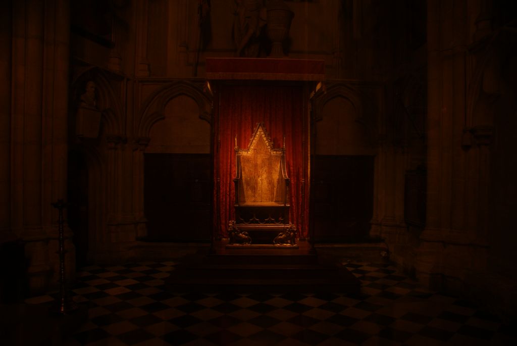 An old gilded wooden throne stands inside a red-curtained display on a black-and-white checkerboard floor in a very dark stone interior.