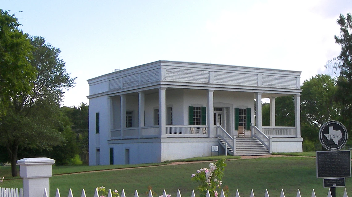 A white columned house with a raised verandah and central steps stands in a green landscape.