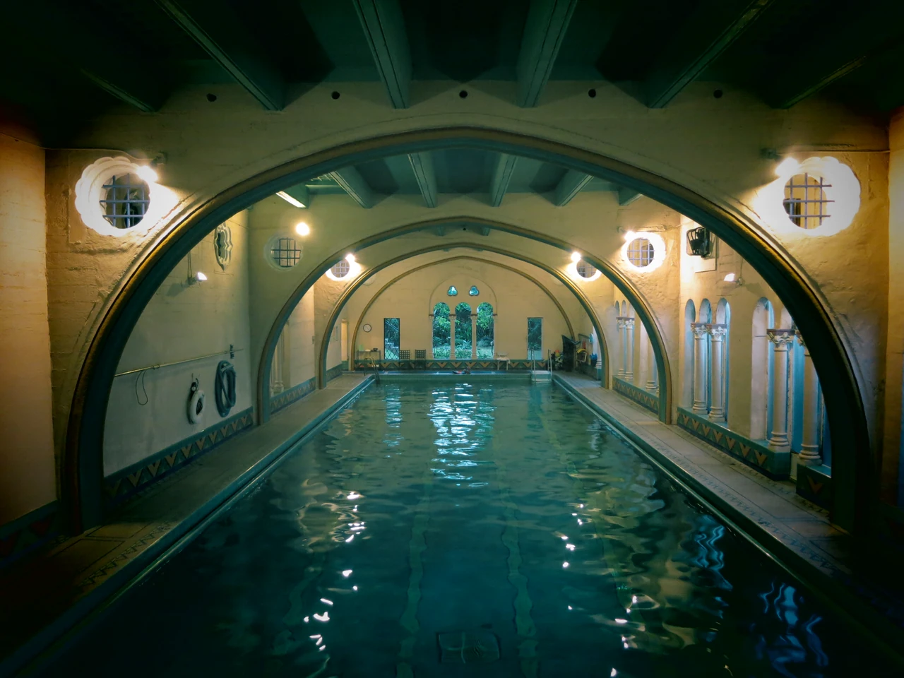 Repeated arches span a long indoor swimming pool, with side columns, small round windows and reflected light on the water.