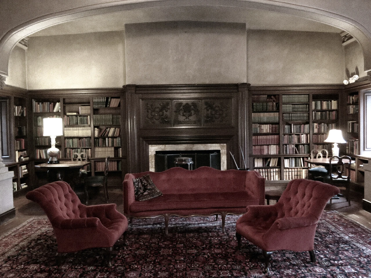 Bookcases and a fireplace surround upholstered seating beneath a pale curved ceiling in a furnished club room.