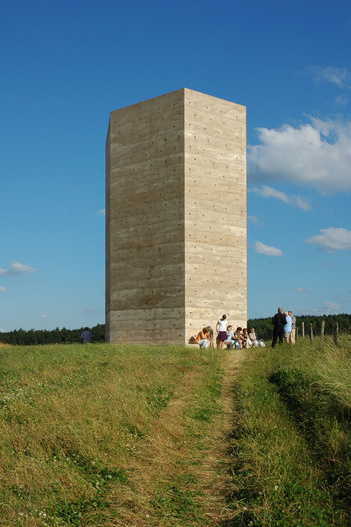 A tall compact chapel with visibly layered concrete walls rises above a grassy field, with visitors at its base.