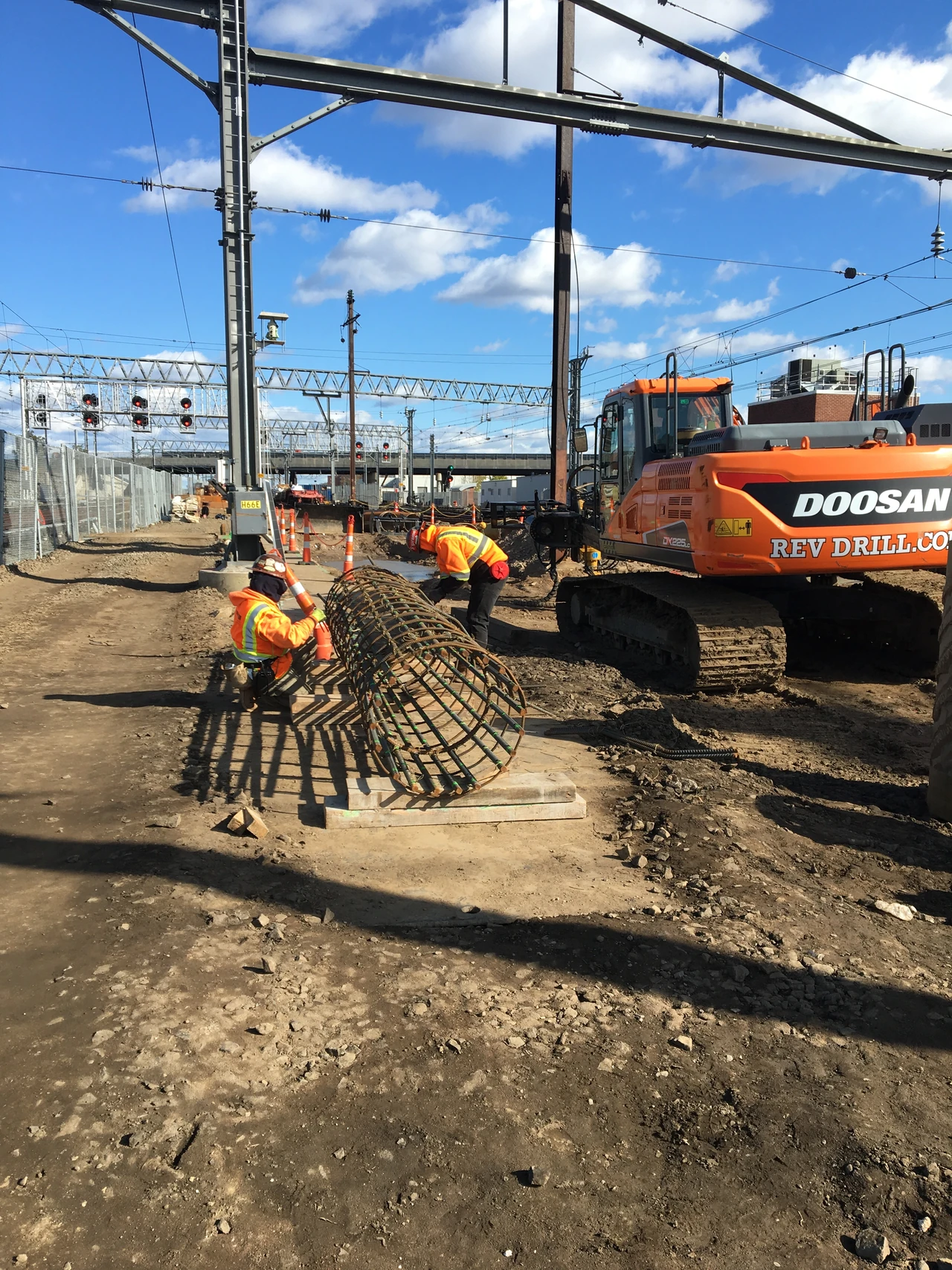 Two workers prepare a cylindrical reinforcement cage beside excavating machinery at a railway construction site.