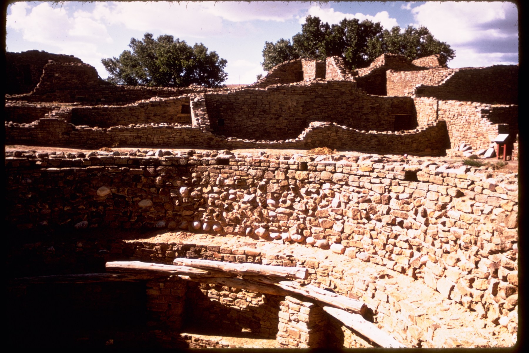 Low rough-stone walls and coursed stone rooms with door openings and a timber across the foreground at Aztec Ruins.