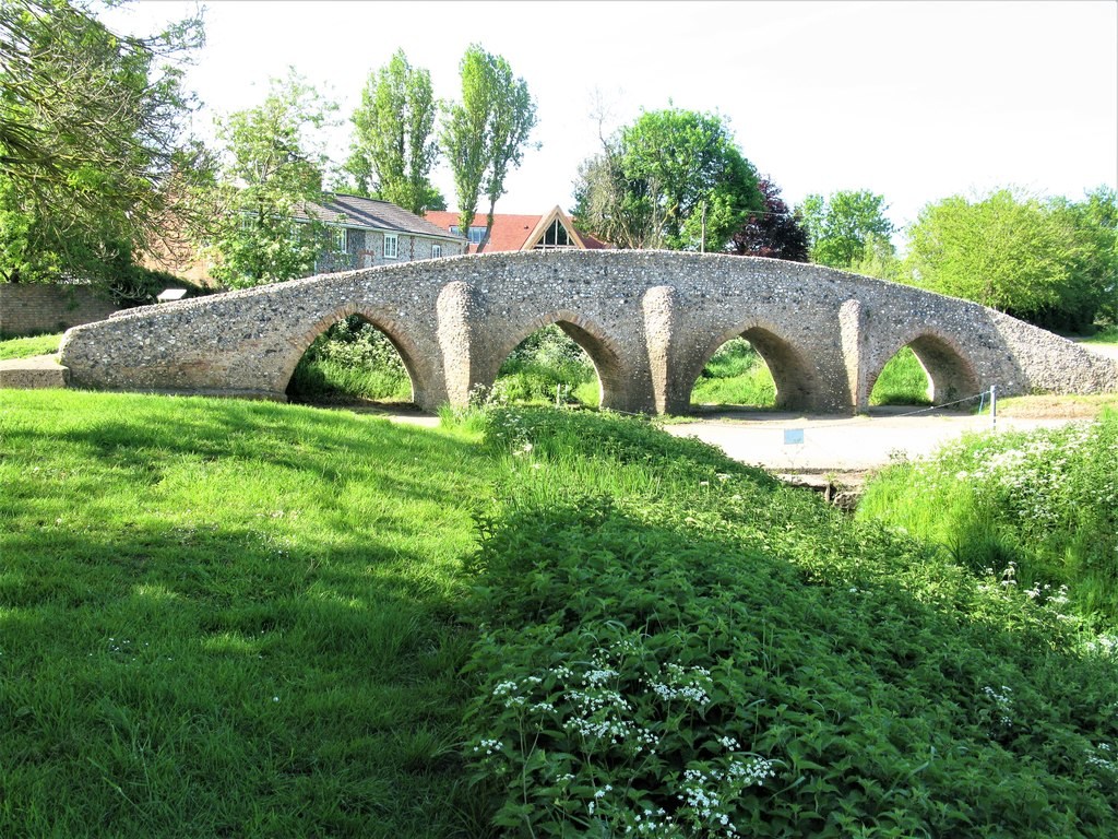 Flint-faced Moulton Packhorse Bridge with four brick-lined arches, projecting piers and grassy banks.