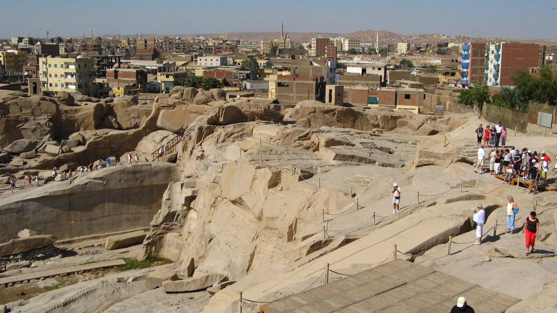 An unfinished obelisk in the exposed granite quarry at Aswan, with visitors and the city behind.