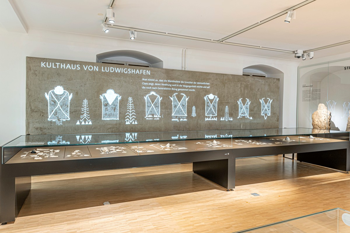 Ancient clay wall fragments in glazed museum cases stand in front of a modern projection of reconstructed figures.