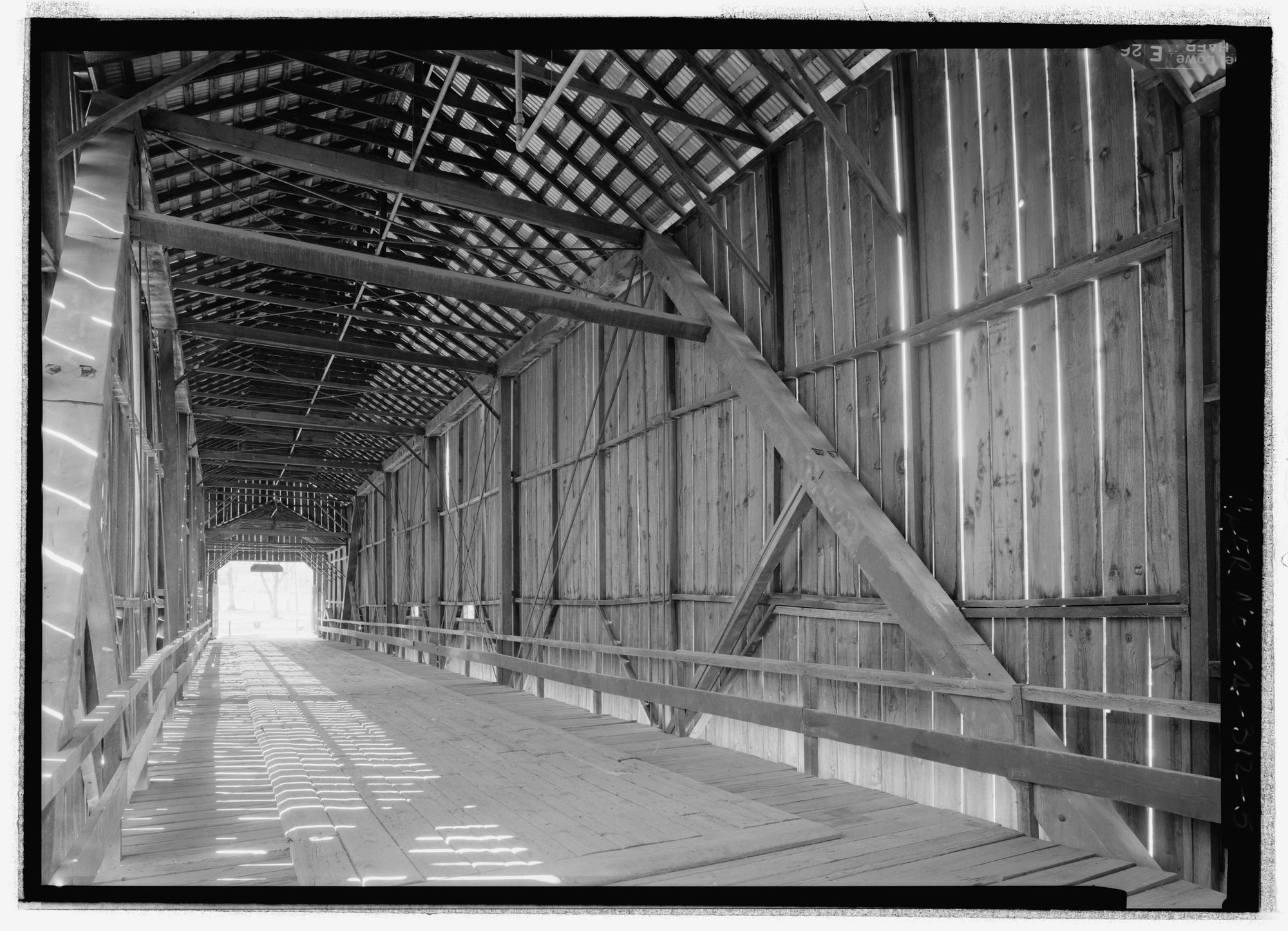 Greyscale view down the covered bridge interior, showing diagonal timbers, decking and daylight beyond.