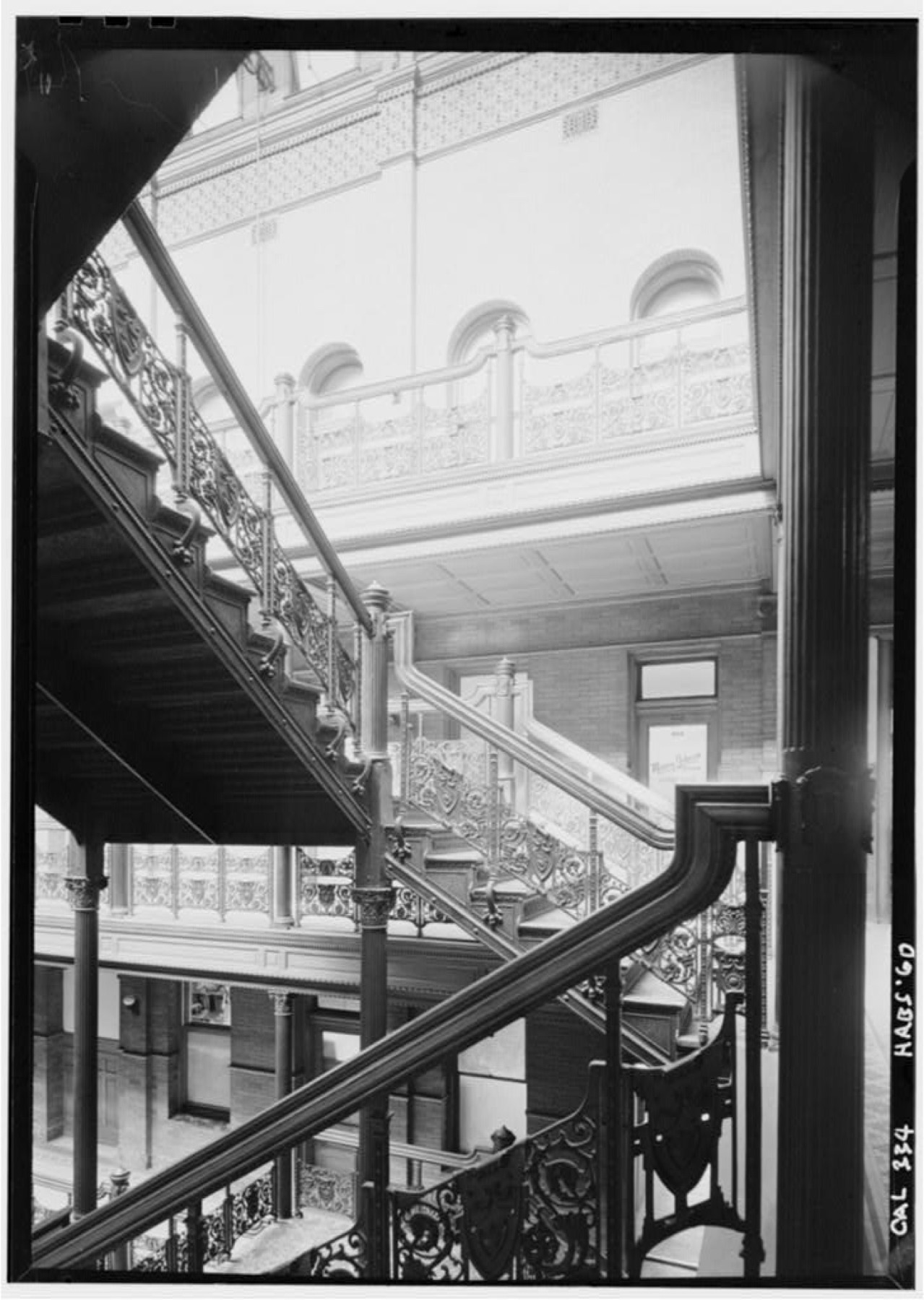 Black-and-white view along Bradbury’s glazed internal court with repeated metal balconies, stairs and bright roof light.