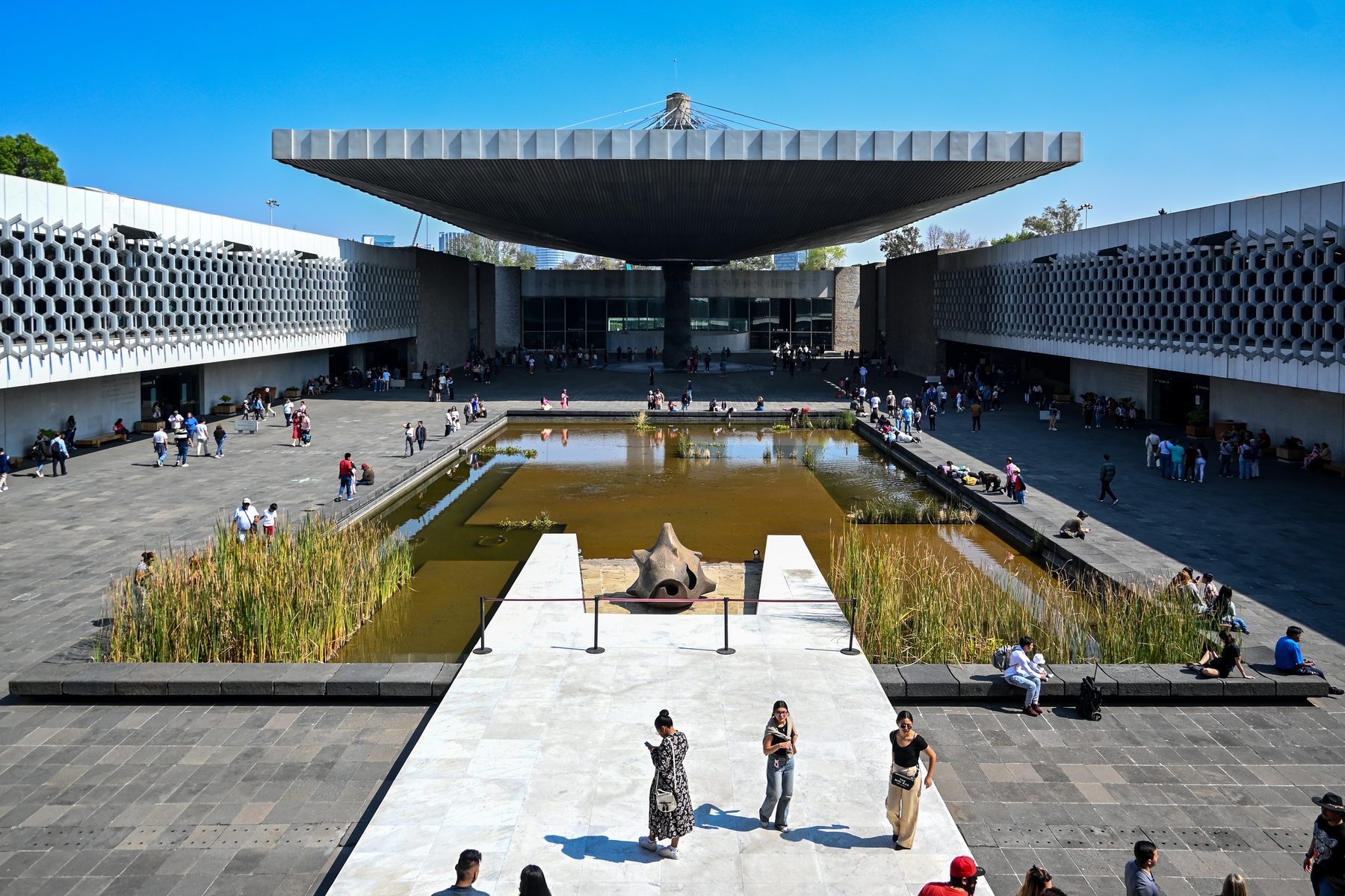 Museum courtyard seen toward the large partial umbrella canopy with visitors, pond and surrounding gallery edges.