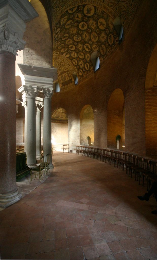 A tiled passage curves between paired columns and a brick wall with niches, beneath a mosaic-covered barrel vault.