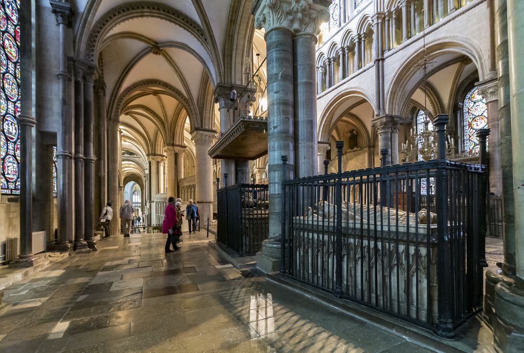 Visitors stand on a vaulted passage beside tall windows and an iron enclosure containing a tomb.