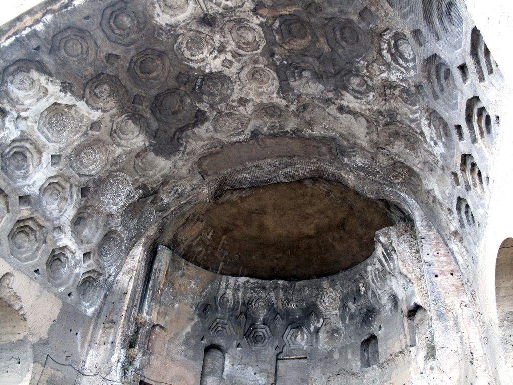 A curved wall with niches and a semi-dome is recessed beneath a much larger coffered barrel vault in a Roman ruin.