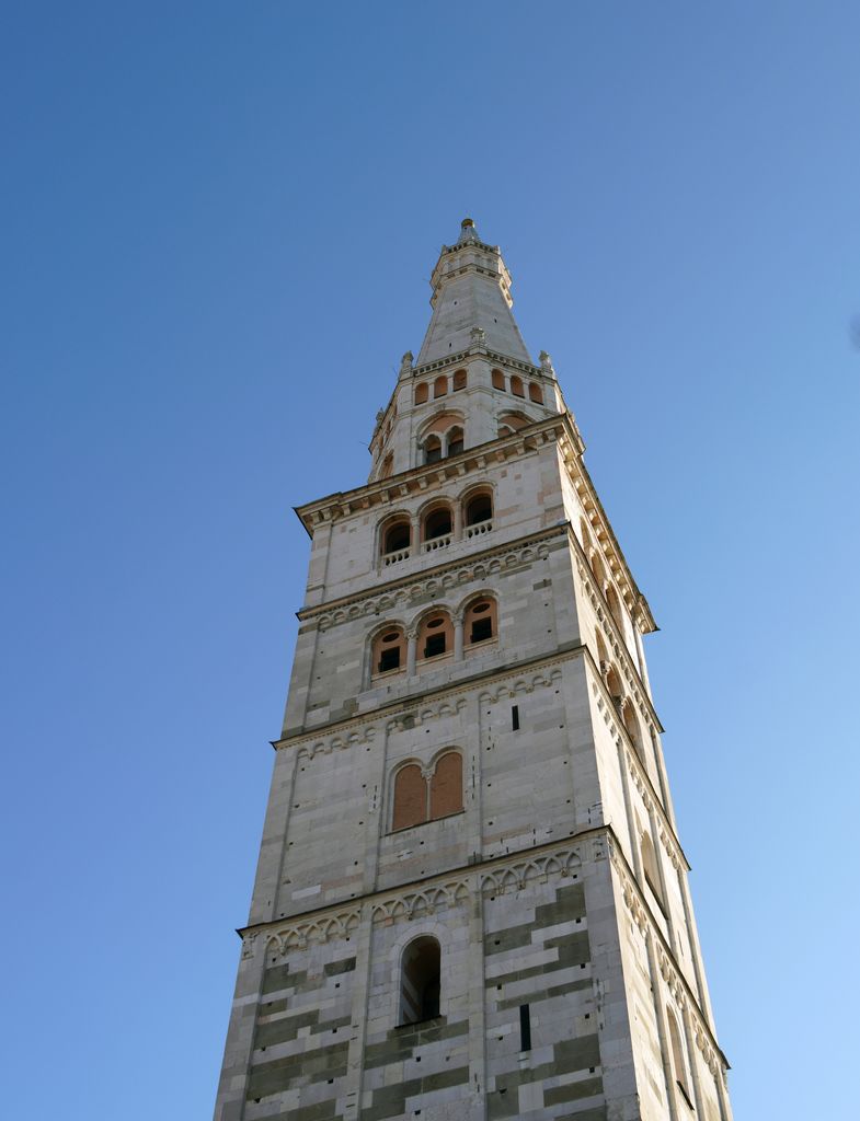Two pale masonry faces with repeated arched openings rise into a narrower polygonal upper section and pointed spire.