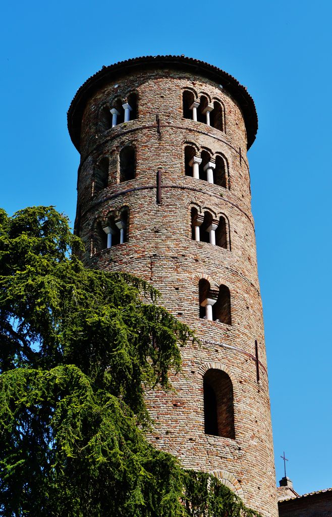 The curving brick tower has single, paired and triple arched openings beneath a low circular roof, with foliage concealing part of its lower side.