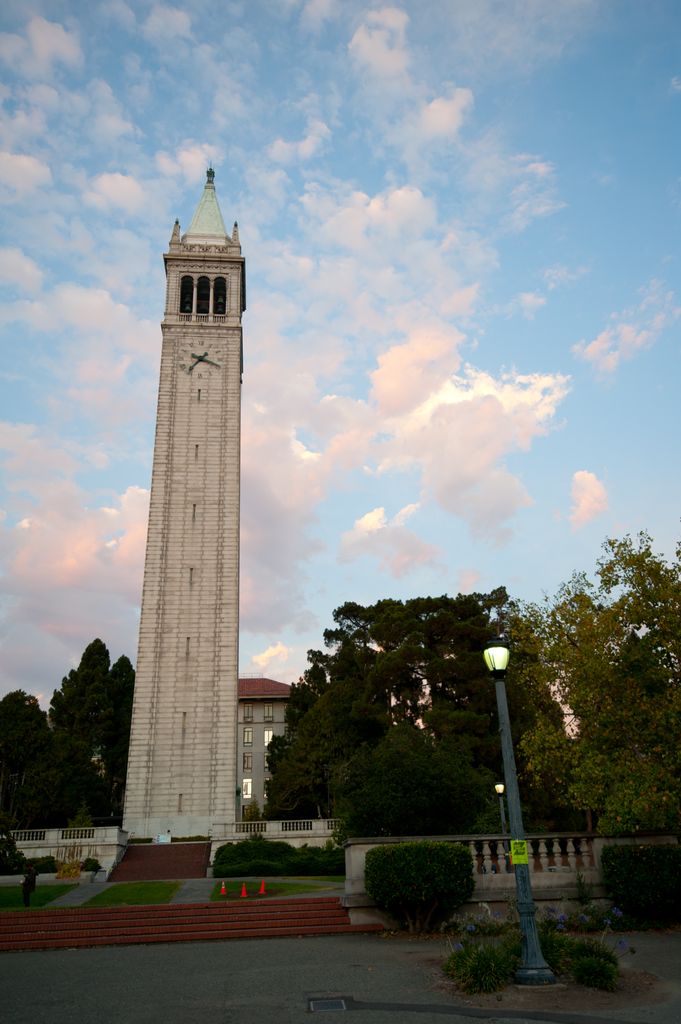 A freestanding pale clock and bell tower rises above campus steps, terraces and trees beneath evening clouds.
