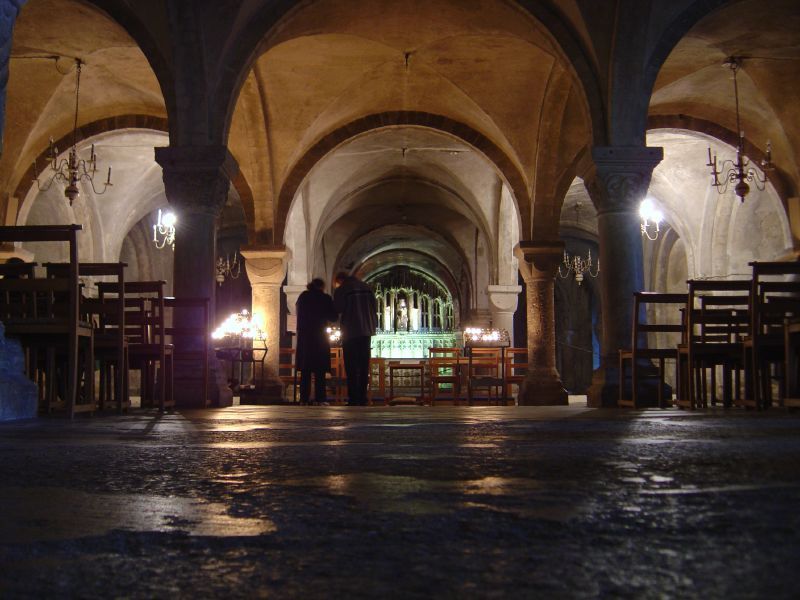 Low rounded arches and stone vaults recede between thick supports toward an illuminated altar area, with chairs and two visitors.
