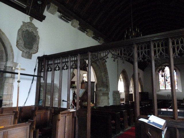 A dark timber screen with traceried openings divides a church interior beneath a continuous roof, with seating and masonry arches visible through it.