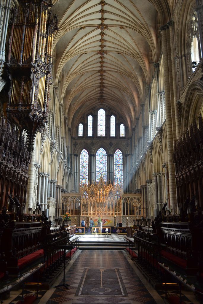 Dark facing stalls frame a central passage toward steps, a brighter open altar area and a reredos beneath a high windowed vault.