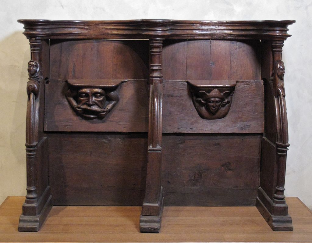 Two adjoining wooden stalls have raised hinged seats, each exposing a carved face beneath a small projecting support.