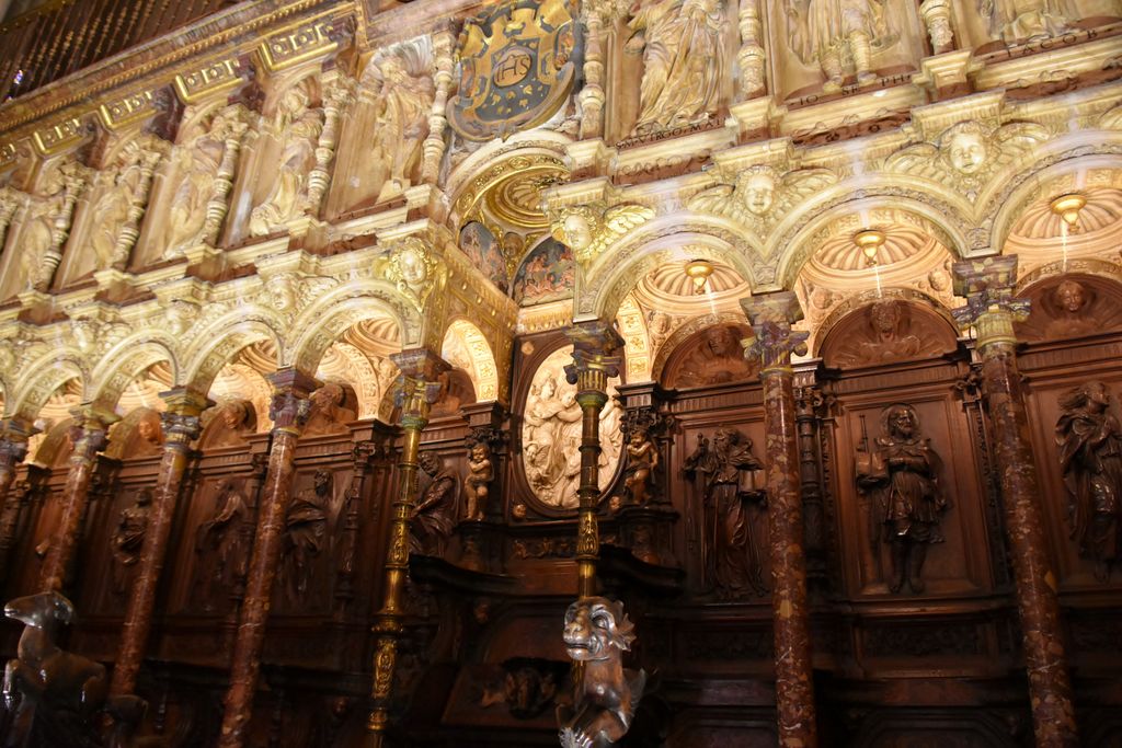 Dark wooden seats and carved backs stand beneath a lighter sculpted arcade, with decorative figures and varied stonework above.
