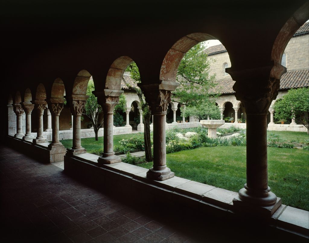 Carved capitals and stone columns frame a planted court and fountain, viewed from a shaded covered gallery.