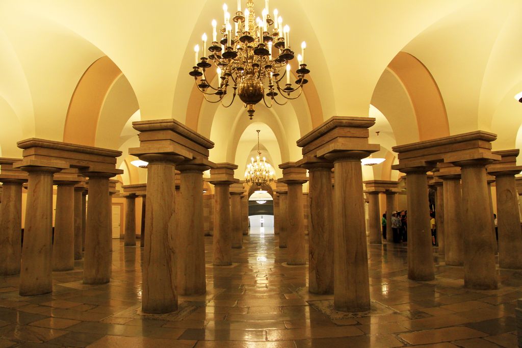 Many stout Doric columns support arches and pale vaulting above a paved civic interior lit by chandeliers.