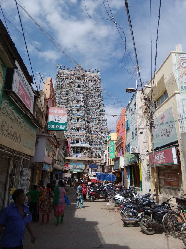 A tall painted temple gateway rises above a narrow street lined with shops, motorcycles, wires and pedestrians.