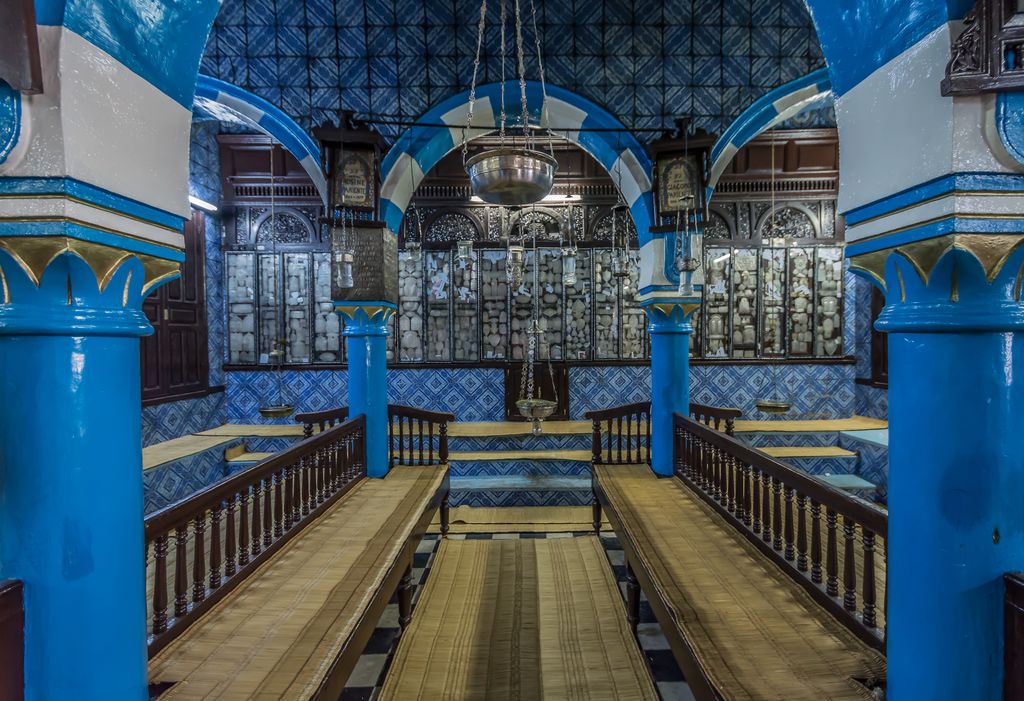 Blue columns and striped arches frame glass-fronted Torah-ark cabinets beyond wooden benches and a narrow central passage.