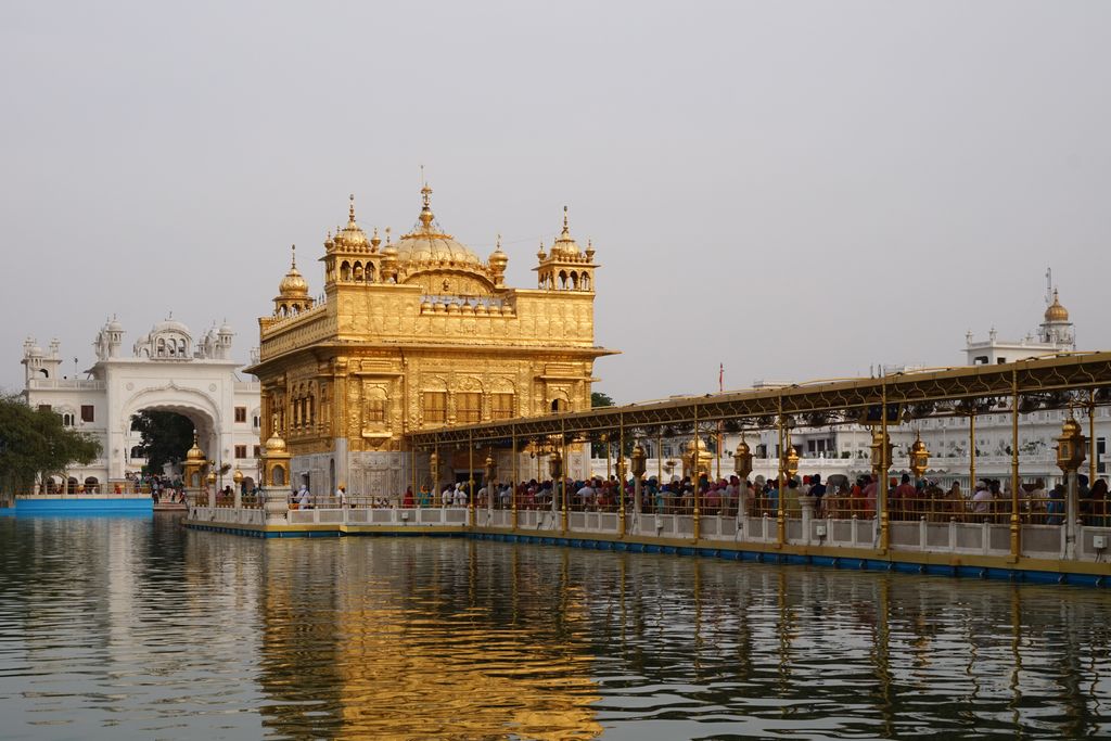 A covered walkway crowded with visitors crosses the pool from the right toward a gilded shrine reflected in the water.