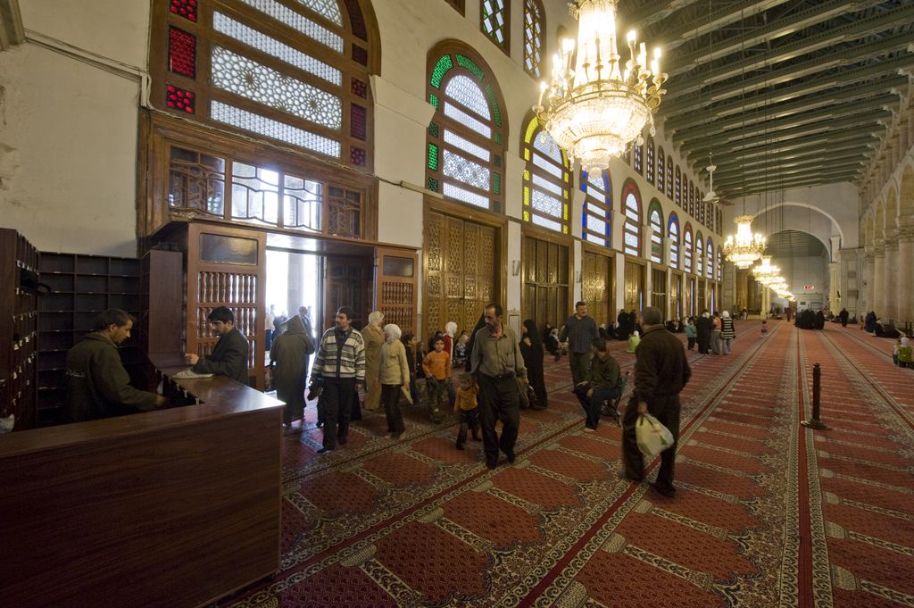 Visitors gather beside tall doorways and stained-glass windows in a long carpeted aisle beneath repeated ceiling beams and chandeliers.